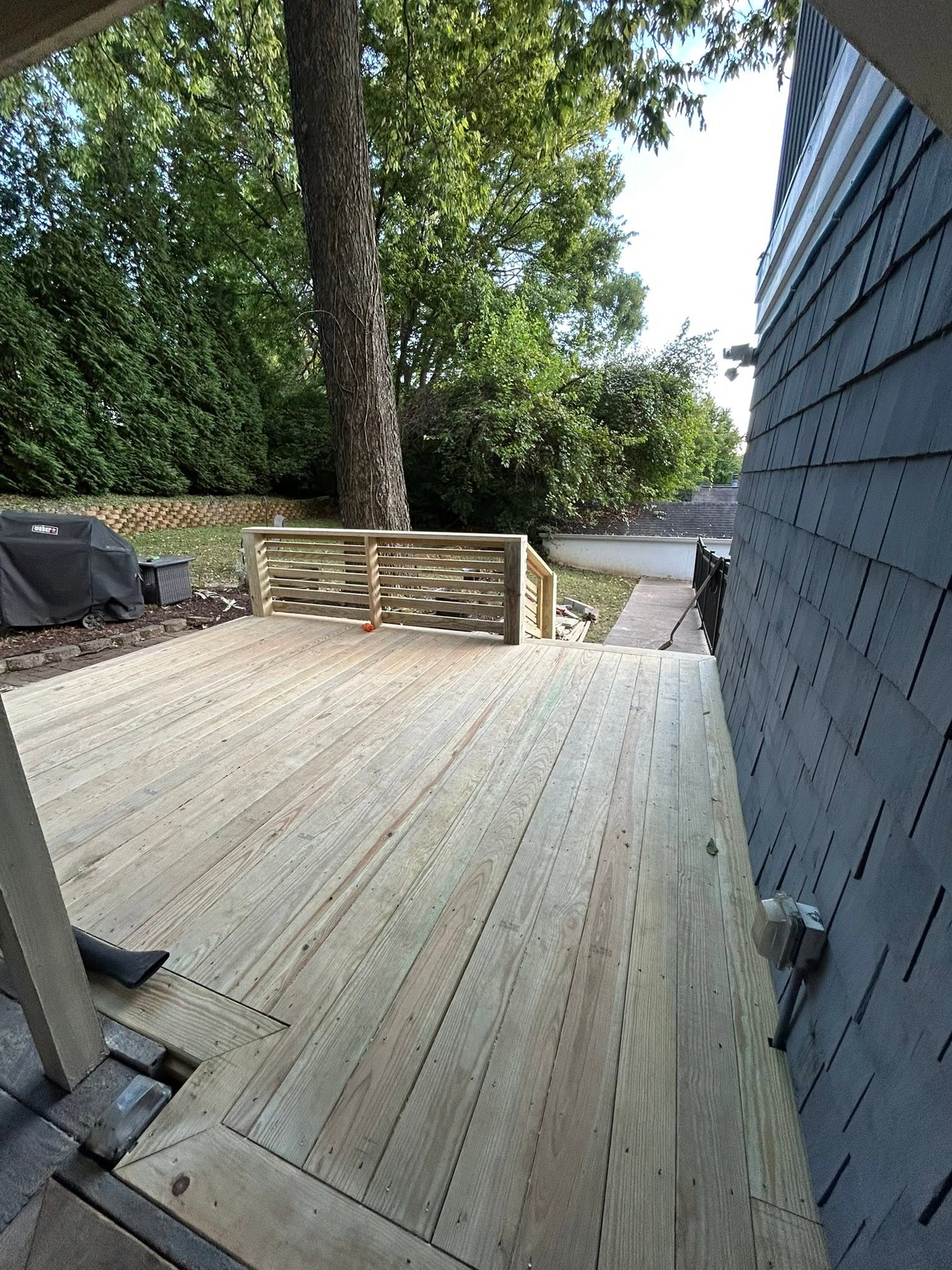 Wide wood deck with horizontal-slat railing beside a blue-sided home and leafy backyard