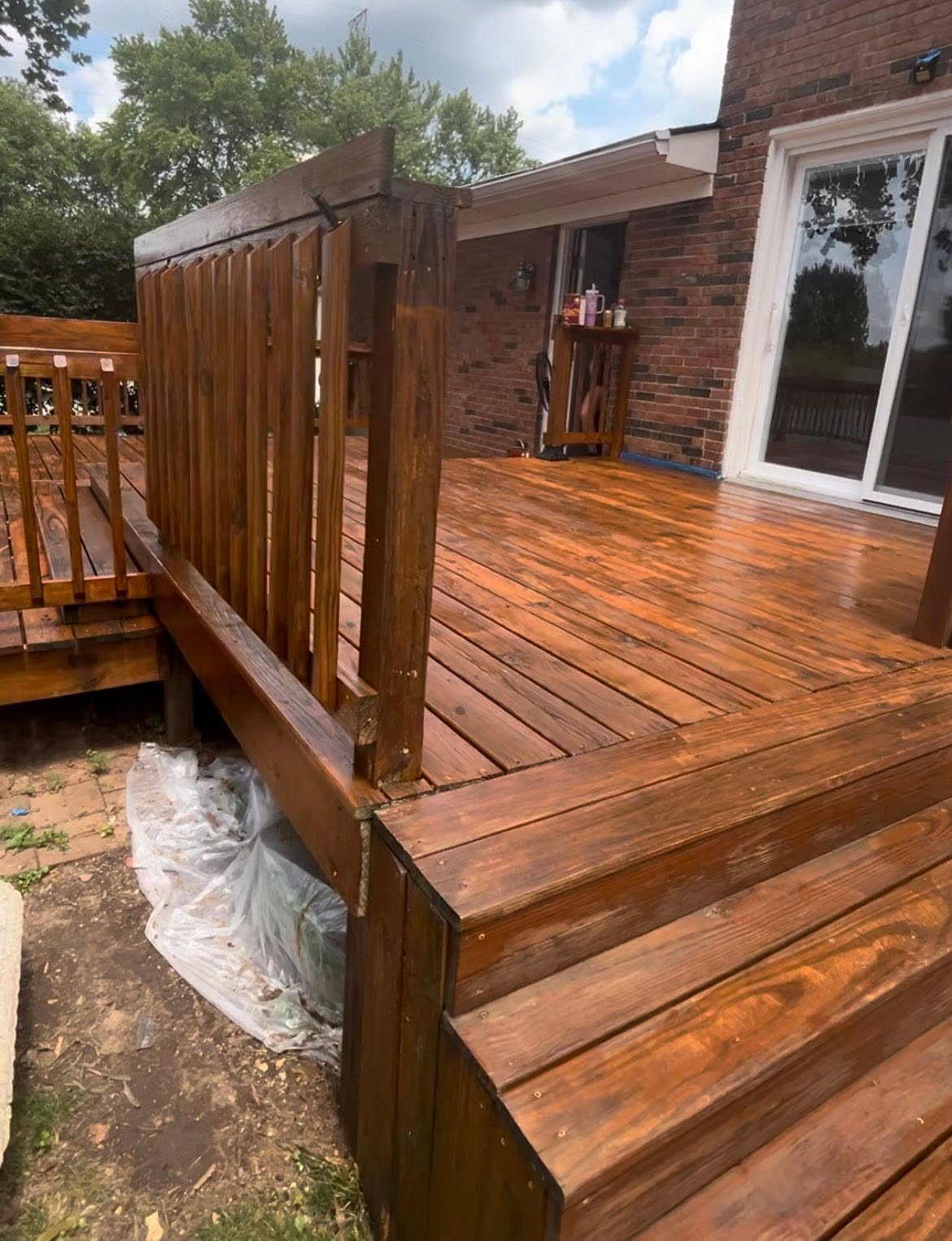 Warm brown wood deck, railings and steps beside a brick home, with protective plastic beneath the edge